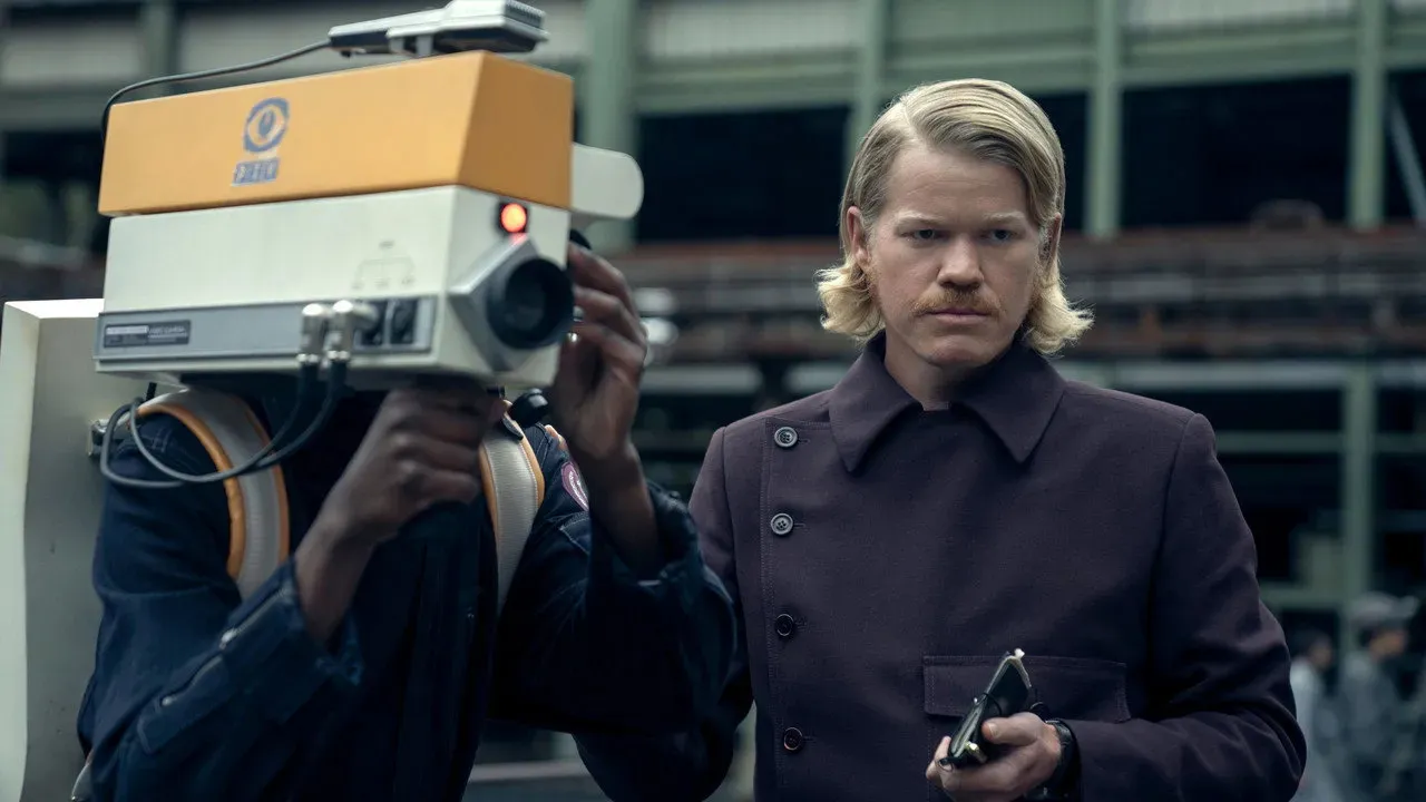 Jesse Plemons as Plutarch Heavensbee, standing beside a Capitol camera operator in Sunrise on the Reaping